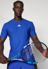 Modra rebrasta športna majica s kratkimi rokavi, ki ima belo logo. Oseba drži teniško lopar s črno-modrim vzorcem.