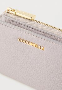 Vaaleanlaventelinvärinen nahkainen vetoketjupussi, jossa on teksturoitu pinta, kultainen vetoketjun metalliosat ja kohokuvioitu "Coccinelle"-logo.