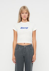 White cropped t-shirt with blue "Roxy" logo, short sleeves, and a minimalist design, paired with gray high-waisted pants.