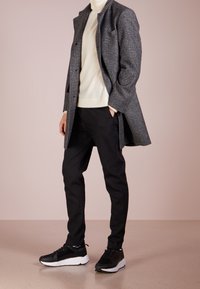 Grå struktureret frakke over en beige trøje, parret med sort slim-fit bukser og sorte sneakers med hvide såler. Glat overflade, skræddersyet look.