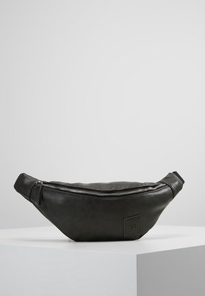 Marsupio in pelle nera con cerniera argentata e una piccola toppa cucita con le iniziali "Y.T." sul davanti, posato su una superficie bianca.