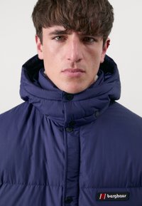 Doudoune bleu marine avec col montant, boutons-pression et patch logo Berghaus. Texture lisse et design matelassé pour l'isolation.