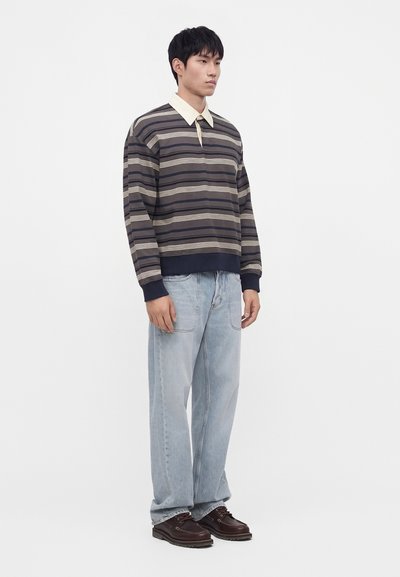 Homme debout portant une chemise rayée à manches longues avec un col beige, un jean bleu clair et des chaussures marron foncé sur un fond uni.