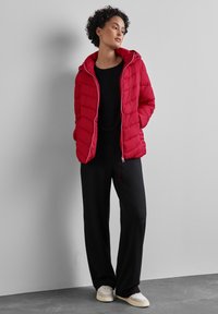 Rote Pufferjacke mit vertikaler Naht, Kapuze und silberfarbenen Akzenten, kombiniert mit einem schwarzen Oberteil und lockeren schwarzen Hosen. Weiße Sneaker runden den Look ab.