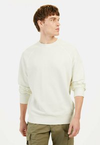 Helle beige Sweatshirt mit Raglanärmeln, Rundhalsausschnitt und gerippten Bündchen. Weiche Textur, minimalistisches Design, kombiniert mit olivfarbenen Cargo-Hosen.