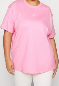 Person som bär en lös rosa Adidas t-shirt och vita byxor, visad från halsen till höfterna mot en enkel bakgrund.