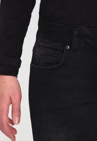 Černé džíny z denimu s přiléhavým střihem, se přední kapsou a viditelnými švy. Materiál má mírný lesk a hladkou texturu.