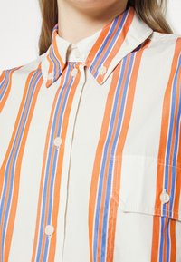 Vit skjorta med vertikala ränder i orange och blått. Har en knappar-ned krage och en bröstficka. Slät tygtextur.