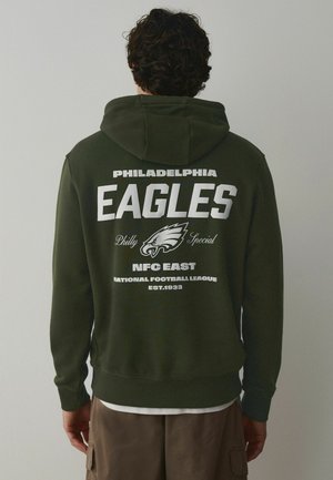 Man wearing dark green hoodie with Philadelphia Eagles logo and text, standing facing a plain light background.