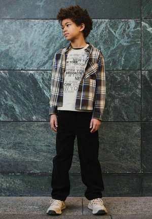 Un garçon aux cheveux bouclés portant un tee-shirt graphique beige de New York, une chemise à carreaux, un pantalon noir et des baskets blanches, se tenant contre un mur en pierre verte.