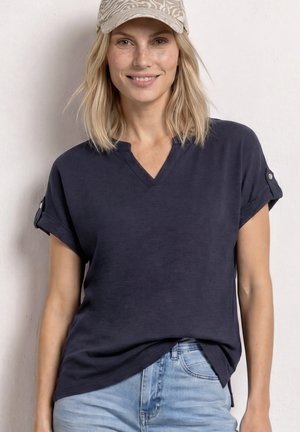 Femme souriante portant une casquette légère à motifs, un haut bleu marine à col en V manches courtes avec des détails de boutons, et un jean bleu clair devant un mur uni.