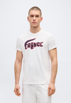 Νεαρός άνδρας με κοντά μαλλιά που φοράει λευκό μπλουζάκι με κόκκινο και μαύρο κείμενο "FRANCE" και σχέδιο ουράς κροκόδειλου σε απλό φόντο.