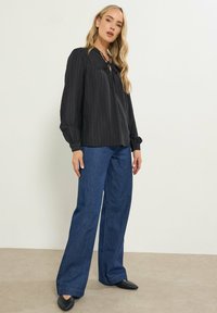 Unge kvinde med langt blont hår iført en sort bluse med lange ærmer, brede blå jeans og sorte spidse sko foran en ensfarvet væg.