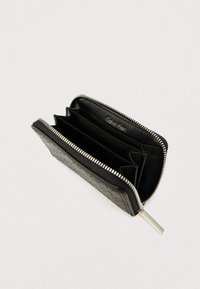 Μαύρο πορτοφόλι από δέρμα με υφή στην επιφάνεια, ασημένιο φερμουάρ και τρία εσωτερικά διαμερίσματα για κάρτες και μετρητά. Εσωτερική ετικέτα με το λογότυπο Calvin Klein.