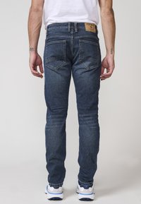 Pantalones vaqueros de mezclilla azul oscura con corte ajustado, que cuentan con bolsillos traseros y una etiqueta marrón en la cinturilla. Llevados con zapatillas blancas y azules.