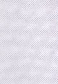 Zbliżenie na białą teksturowaną tkaninę z drobnym, równomiernie rozmieszczonym wzorem dzianinowym tworzącym subtelną powierzchnię przypominającą siatkę.