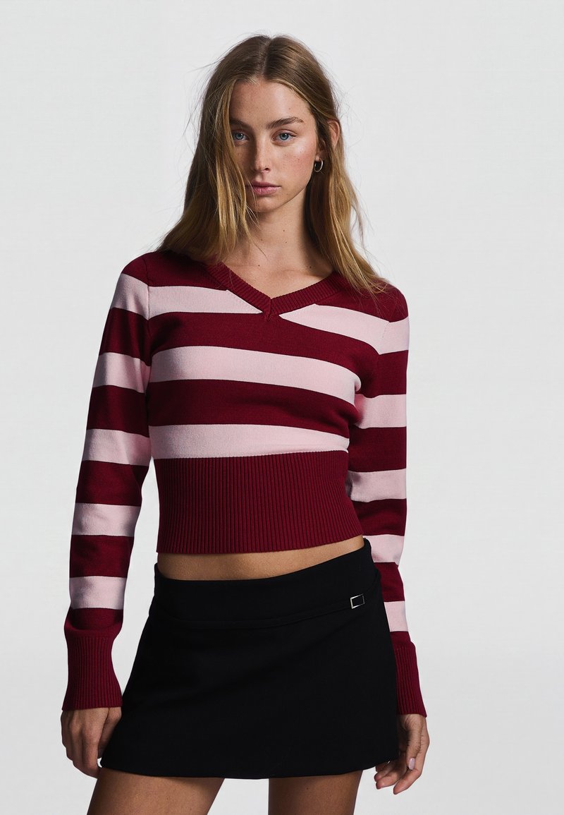 Femme aux longs cheveux blonds portant un pull court rayé bordeaux et rose clair et une mini-jupe noire, debout devant un fond uni.