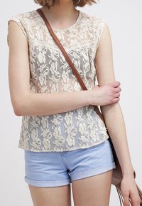 Blusa in pizzo con motivo floreale, colore crema, scollatura rotonda, maniche a sbuffo, abbinata a pantaloni corti di jeans blu chiaro arrotolati e una borsa a tracolla marrone.