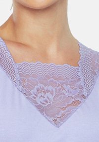Liliowa bluzka z dekoltem w serek, ozdobiona koronką z kwiatowym wzorem. Koronka ma falbaniaste krawędzie oraz fakturowany wzór, co kontrastuje z gładkim materiałem.