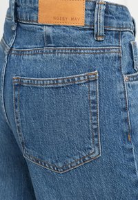 Blugi din denim albastru cu talie înaltă, cusături vizibile și o etichetă din piele inscripționată cu "NOISY MAY" pe centura din spate.