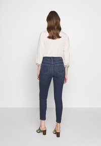 Skinny jeans in dark blue denim with a high waist, featuring back pockets and subtle stitching. Paired with a light shirt and heeled sandals.