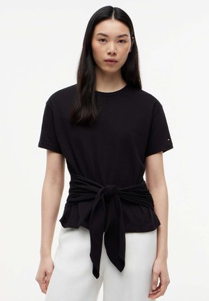 Donna con lunghi capelli neri, indossa una maglietta nera annodata in vita e pantaloni bianchi, in piedi davanti a uno sfondo bianco semplice.