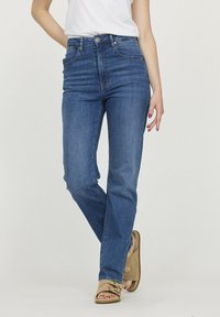 Jeans en denim bleu taille haute avec une coupe ajustée, décoloration subtile, cinq poches et fermeture à bouton. Portés avec des sandales beiges.