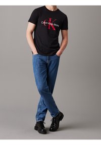 Muž v černém tričku Calvin Klein Jeans, modrých džínech a černých kožených botách, stojící s rukama v kapsách na šedém pozadí.