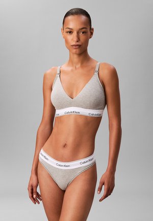 Kobieta ubrana w jasnoszary biustonosz i pasujące do niego majtki Calvin Klein, stojąca na szarym, jednolitym tle, zwrócona przodem.