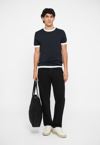 T-shirt azul-escuro com gola e punhos canelados brancos, calças pretas, ténis de cor creme e uma mala tote preta. Design simples e ajuste casual.