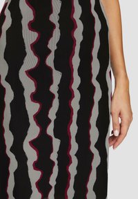 Tissu noir avec des rayures ondulées en gris et bordeaux. La texture semble lisse avec un léger éclat. Une main est visible tenant le tissu.