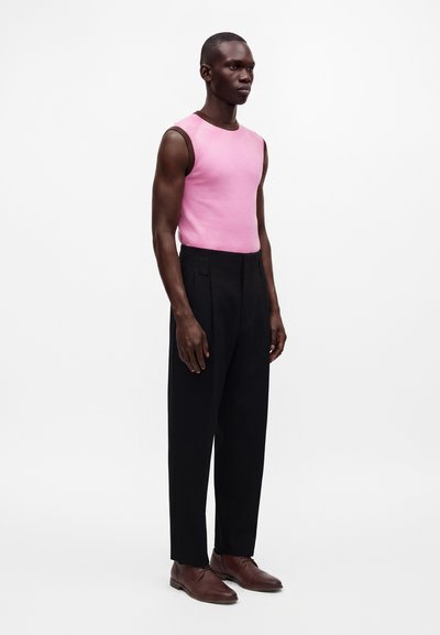 Homme portant un haut rose sans manches, un pantalon noir à jambes larges et des chaussures marron, debout devant un fond blanc uni.