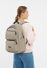 Egy fiatal nő világos rózsaszín pulóvert és kék farmert visel, és egy nagyméretű bézs hátizsákot cipel, amelyen több cipzáras zseb található.