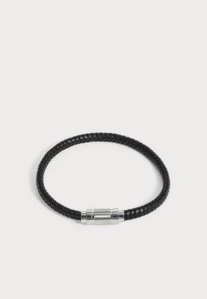 Pulsera de cuero trenzado negro con cierre magnético cilíndrico plateado sobre un fondo liso y claro.