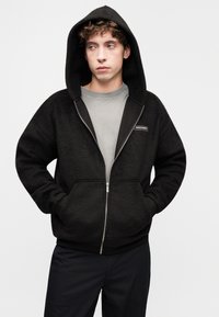 Sweat à capuche noir zippé en tissu doux et texturé. Comprend une poche frontale, une capuche réglable et un léger logo sur la poitrine.