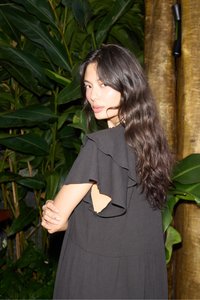Jeune femme aux longs cheveux ondulés et foncés portant une robe noire à volants, debout de profil, les bras croisés devant un fond de grandes feuilles vertes.