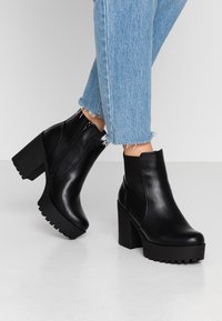 Mustat nahkaiset nilkkurit paksulla korolla ja kumipohjalla, joissa on vetskari ja sileä tekstuuri. Rennot kulutetut farkut.