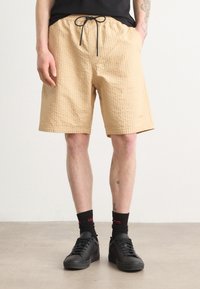 Beige quilted shorts with an elastic waistband, black drawstring, and side pockets, paired with black sneakers and dark socks.