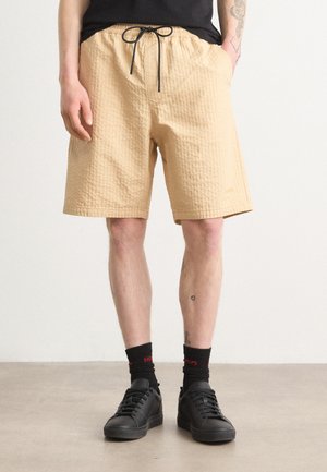 Beige quilted shorts with an elastic waistband, black drawstring, and side pockets, paired with black sneakers and dark socks.