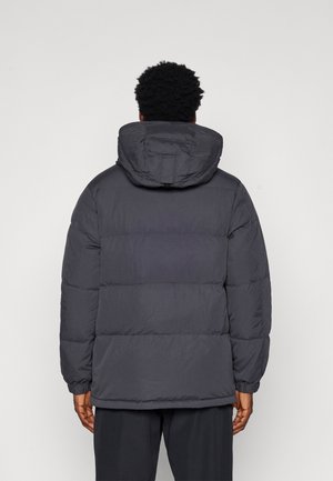 Veste grise matelassée avec une capuche, présentant un design matelassé, des manches longues et des poignets élastiques. La texture semble lisse et isolante.