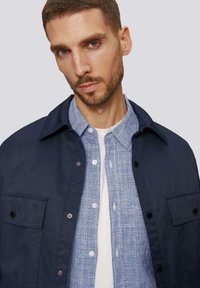 Marineblauwe button-up jas over een lichtblauw geruit overhemd. De jas heeft een zak en zwarte drukknoopjes. Het overhemd heeft een geweven textuur.