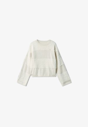 Jeune fille aux longs cheveux porte un pull blanc oversize, une jupe en denim bleue, des chaussettes beiges et des chaussures noires, debout devant un fond gris.
