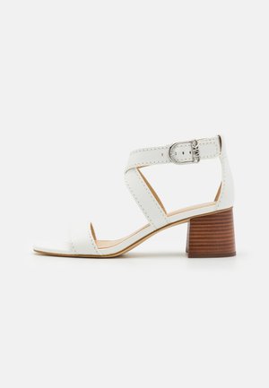 Sandale blanche en cuir à bout ouvert, bride cheville, boucle argentée et talon moyen en bloc de bois empilé sur fond uni.