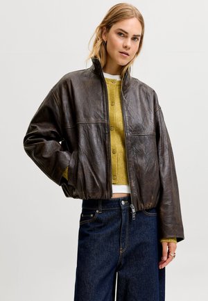 Femme portant une veste en cuir marron foncé par-dessus un cardigan jaune boutonné et un jean bleu foncé, debout avec une main dans la poche de la veste.