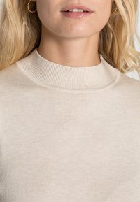 Pulover tricotat bej cu guler înalt, având o căptușeală texturată și o suprafață netedă. Viziunea de aproape subliniază detaliile cusăturii.