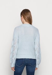Ljusblå stickad tröja med ribbad nederkant och ballongsärmar som har ett öppet vävmönster. Bakifrån syns texturerad detalj på ärmarna.