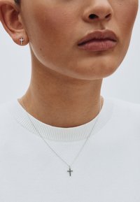 Strieborné náušnice v tvare kríža a zodpovedajúci náhrdelník, oba s minimalistickým dizajnom, umiestnené na pozadí bieleho svetra.