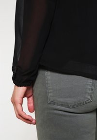 Czarna, przezroczysta bluzka z marszczonymi mankietami, zestawiona z szarymi, dopasowanymi spodniami z tylną kieszenią. Skup się na fakturze tkaniny i dopasowaniu.