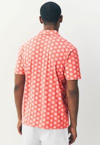 Camisa de manga corta en tejido color coral con un patrón repetitivo de rayos de sol en blanco. Cuello clásico y textura suave.