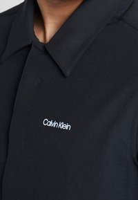 Sort polo-shirt lavet af blødt stof med klassisk krave, knaplukning og broderet hvid "Calvin Klein" logo på brystet.